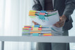 © Kritdanai - Organized Office Task: A person organizes a stack of paperwork, neatly arranged with colorful tabs and clips, against the backdrop of a bright, minimalist office setting.