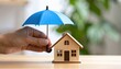 © Igor_Tichonow - Hand shielding wooden house with umbrella symbolizing home insurance, protection from risk, safety planning and financial stability for property owners