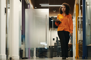 Wall Mural - Young Black woman walking through modern office hallway holding digital tablet, looking ahead with focused expression, business environment visible in background