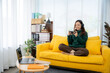 © Kritdanai - Cozy Gamer Girl in Living Room: A young woman immersed in a mobile game, radiating tranquility while perched comfortably on a yellow sofa in a modern living room.