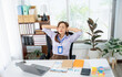 © Kritdanai - Exhausted At The Desk: A portrait of a businesswoman with her hands behind her head sitting at her desk with a laptop and documents, displaying a clear indication of stress.