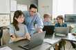 © AnnaStills - Caucasian teenage girl with prosthetic arm sitting at desk using laptop while middle aged teacher standing beside her pointing at screen, two Caucasian teenage boys working on laptops in background