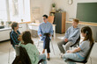 © AnnaStills - Middle aged Caucasian woman leading group therapy session with four teenagers of diverse ethnicities sitting in circle, engaging in discussion in classroom setting, sunlight streaming through window