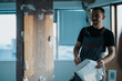 © qunica.com - A person wearing overalls stands in a building under renovation, holding a document and looking focused.