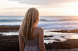 © Pixels Pictures - A serene beach photograph of a young woman with long blonde hair from behind, wearing a light gray sleeveless dress, standing on rocky volcanic terrain.