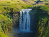 Skogafoss waterfall in Iceland cascades from a high cliff, surrounded by green hills and rugged terrain. Mist rises at the base, with people visible nearby.