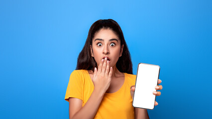 Portrait of emotional excited and surprised young indian woman holding smartphone with black blank screen in hand, showing device, covering mouth with hand. Gadget with empty free space for mock up