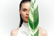 © VICHIZH - Young woman with beautiful skin partially hidden by a tropical leaf, showcasing natural beauty and serenity in a minimalist setting.