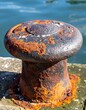 © AsterionMad - Close Up of Rusted Iron Bollard on a Dock in Bright Sunlight with Sea in Background