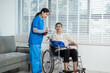© NanSan - A young Asian nurse attentively takes notes while conversing with an elderly woman in a wheelchair with an arm sling,calmly seated in a bright living room receiving medical and rehabilitative care