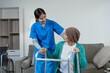© NanSan - A young Asian nurse assists an elderly female cancer patient, wearing a headscarf, with using a walker. The patient sits on a sofa, preparing for rehabilitation and mobility training in a caring