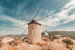 © EdNurg - Traveler admiring a historic stone windmill under a bright sky, surrounded by a picturesque landscape of hills and clouds