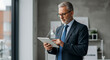 © Rsquare stock - mature older business man executive standing in office using digital tablet. Middle aged professional businessman corporate manager wearing suit and glasses holding tab working on finance project