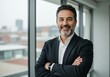 © sujon1638 - Confident middle aged businessman with salt and pepper beard and arms crossed smiling warmly by a large window with cityscape view