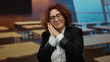 © Krakenimages.com - Woman stretching and gesturing sleepily in a classroom with wooden desks and modern design, wearing glasses and a suit, middle-aged hispanic brunette in an educational setting.