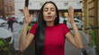© Krakenimages.com - Young hispanic woman in a red shirt meditates with closed eyes on a busy city street, surrounded by cars and urban architecture, conveying peace amid urban chaos.
