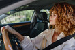 © SHOTPRIME STUDIO - Curly woman driving a car with focused expression, wearing a seatbelt in a modern vehicle interior, natural daylight, spring season outside the window.