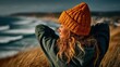 © MO - Woman in a yellow beanie and green hoodie looking out at the ocean, hands behind head, enjoying the beach scene.