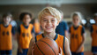 © Julia Zarubina - Smiling blond boy 10 years old in basketball uniform holding basketball ball in the center of school gym, other children in sports uniform behind, physical education lesson, game, team, training