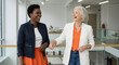© Karmiyem - Two successful businesswomen laugh together in a modern office building hallway, showcasing diversity and collaboration in the workplace.