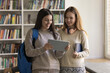 © fizkes - Modern student youth. Confident teen girl gen z learner stand with female groupmate in campus library show new education app on digital tablet share homework answers check test results academic scores