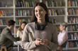 © fizkes - Modern education vibes. Smiling confident gen z female student pose for portrait in public library look at camera hold phone paper book wear earphones on neck use gadgets for learning and social life
