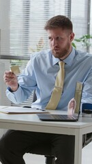 Wall Mural - Vertical shot of young Caucasian male bank agent holding credit card consulting client sitting by laptop at office desk