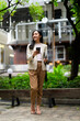 © Parichat - Young Asian businesswoman using smartphone and holding laptop in green urban business district
