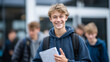 © Maksym - Laughing teenage student showing notes from a notebook to a group of friends, holding the book with one hand while gesturing, school entrance and other students blurred behind