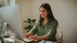 © Hanna Ohnivenko - Woman with long brown hair and warm smile wearing a vibrant green shirt sitting in front of a computer screen with a gentle glow, surrounded by sunlight pouring in through a window to her left, castin