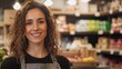 © An - A smiling woman with curly hair standing in a grocery store, wearing an apron, surrounded by various grocery items.