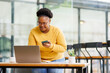 © David - Happy attractive African American business lady using Internet technology for job in co-working space, sitting at laptop, holding mobile phone.