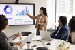 © fizkes - Modern business presentation. Young businesswoman speak to multiethnic professionals group in boardroom meeting explain b2b sales strategy present project stats in charts graphs on digital whiteboard