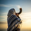 © Mila Shnobel - Person in tallit blows shofar at sunrise, celebrating Rosh Hashanah under a clear blue sky
