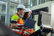 © VStudio - Two people wearing safety gear and holding a laptop. Team of diverse engineers, male and female, programming and operating a CNC machine in a modern factory.
