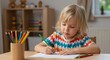 © Tatiane - Blonde child around 6 years old, wearing a colorful shirt, drawing with colored pencils on white paper at a light wooden table. In the background, shelves with toys and soft light entering through a s