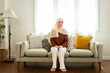 © eakgrungenerd - Muslim woman smile wear hijab sitting on sofa in living room bright, featuring soft lighting and decorative cushions relaxing day off, creating cozy ambiance for relaxation.