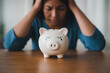 © Paintbrush - A crying piggy bank sits on a table as a worried woman in a blue shirt holds her head.