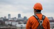 © Helen - Man, a construction worker, wears an orange hard hat and safety harness, looking out over a city skyline. Rooftop view for development.