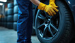 © Viktor - Mechanic in blue uniform and yellow gloves installs new tire onto car wheel. Garage setting with stacked tires in background. Professional auto service, vehicle maintenance and repair.
