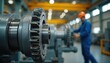 © Viktor - Heavy industrial machinery gear closeup, showing metallic teeth, components in factory setting. Worker in blue overalls, orange hard hat operates machinery in blurred background, suggesting