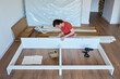 © Lazy_Bear - Woman sits on wooden floor and assembling furniture in new home, fastens parts of white bed frame. DIY home renovation and home organization after moving concept