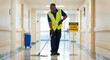 © Thanura - A  janitor mopping a shiny corridor floor in a hospital or public building wearing gloves and safety vest