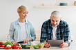 © Prostock-studio - Cook homemade food, recipe app and surfing in internet. Smiling adult lady cuts vegetables, man looks in tablet in kitchen interior with bright vegetables on table, panorama