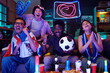 © pressmaster - Group of diverse young adults cheering and celebrating while watching soccer match on television, Black man holding soccer ball, excitement visible on faces, neon lights in background