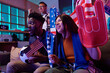© pressmaster - Young Black man holding soccer ball and small American flag sitting next to young Asian woman cheering with foam hand while watching sports event, excited middle aged Caucasian man standing behind