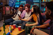 © pressmaster - Group of multiethnic young adults sitting on couch watching smartphone screen, smiling and cheering, celebrating together with drinks and snacks during casual indoor gathering