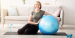 © Prostock-studio - Full length portrait of happy mature woman resting on yoga mat with fitness ball, smiling at camera indoors, free space. Cheerful senior lady taking break from her domestic training