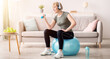 © Prostock-studio - Happy senior woman working out with dumbbells on fitball and listeing to music at home, empty space. Positive mature lady doing strength exercises, keeping in good shape during covid quarantine
