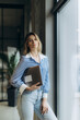 © Roman - Young Casual Woman Holding Laptop Standing by a Large Window